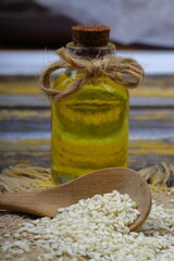 Glass bottle of sesame oil and raw sesame seeds in wooden spoon or bowl on rustic table. Uncooked...