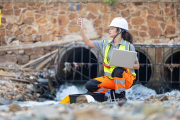 Scientist or Technician Monitoring Water Pollution Near Drain Pipes, Environmental Engineer Testing Water Quality by Culvert, Female Inspector Analyzing Data on Laptop at Urban Waterway