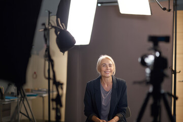 Caucasian mature businesswoman giving a financial report on camera in a recording setup inside her office, lights on, microphone clipped, and branded backdrop behind.