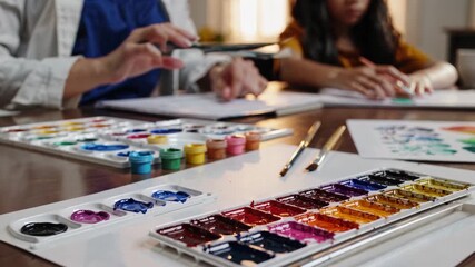 Art teacher picking up a paintbrush from a colorful watercolor palette while teaching a young girl how to paint, sitting at a wooden table with brushes, colorful paint tubes and sheets of paper - Powered by Adobe