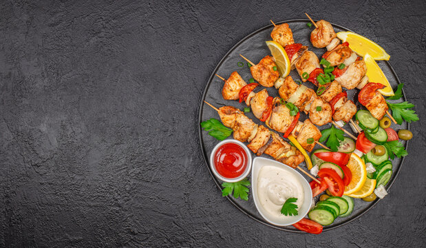 Grilled chicken skewers with fresh vegetables, lemon wedges, and dipping sauces on a dark textured background. copy space, top view