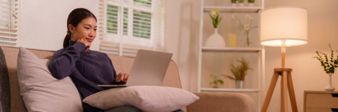 Asian adult woman working from home using laptop computer comfy living room sofa remote online digital lifestyle productivity`