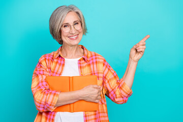 Smiling mature woman holding orange folder against teal background while pointing to the side with...