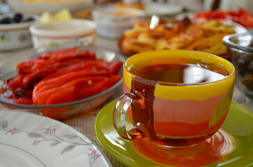A cup of tea sits on a green and yellow plate with a leaf design. The plate is surrounded by a variety of food, including a bowl of peppers and a plate of waffles. Concept of warmth and comfort