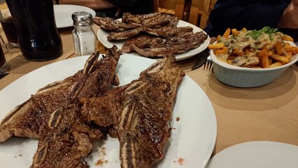 Generous portion of grilled beef short ribs on a plate, with sliced steak and french fries with creamy sauce in the background. A hearty meal served at a steakhouse or traditional uruguayan barbecue.