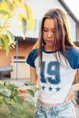 Asian teenage girl in sporty t shirt standing by green tree branches in backyard garden in front of...