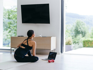 A young woman in sportswear is sitting in the living room and preparing for online training while...