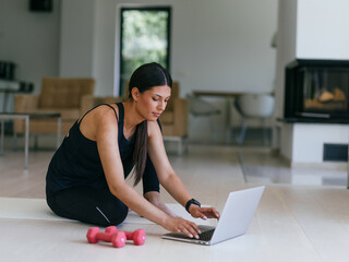 A young woman in sportswear is sitting in the living room and preparing for online training while...