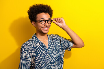 Cheerful young man in a patterned shirt adjusts glasses against vibrant yellow backdrop, expressing...