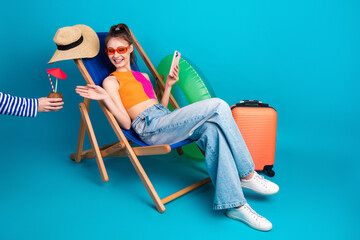 Cheerful young woman seated on a deck chair, holding a phone, with vibrant props, depicting a summer travel moment