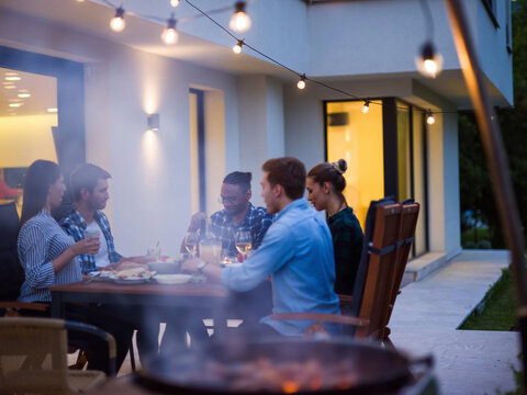 A group of young diverse people having dinner on the terrace of a modern house in the evening. Fun for friends and family. Celebration of holidays, weddings with barbecue