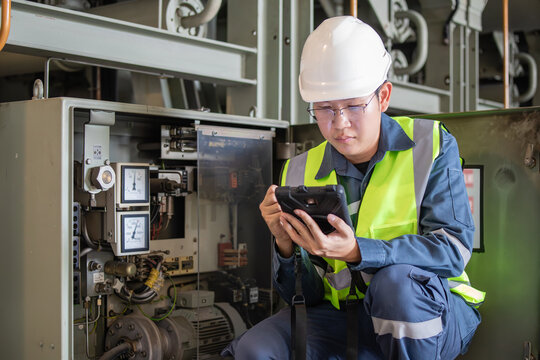 Engineer wearing safety helmet and reflective vest inspects gas insulated switchgear equipment at power station using digital tablet for accurate monitoring and maintenance