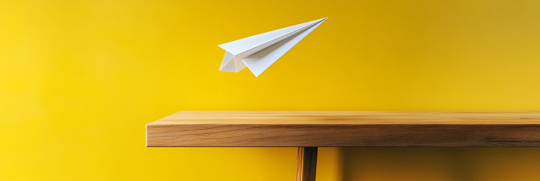 Paper airplane soars above a sleek wooden table against a vibrant yellow backdrop, symbolizing ideas and travel with a minimalist aesthetic. - Powered by Adobe