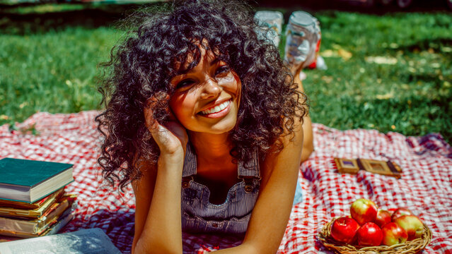 A joyful biracial girl with natural curly hair relaxes on a vibrant picnic blanket in a sunny park