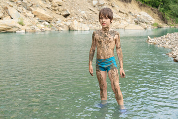 A boy covered in river silt stands in the water of a mountain river
