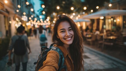 Selfie of smiling woman with backpack walking down street with blurred lights and people behind.