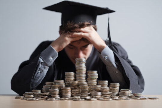 High university tuition fees, unaffordable higher education, the concept. A sad graduate in a gown and master's cap, hunched over a small stack of coins, as debts destroy the graduate's budget