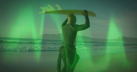 Holding yellow surfboard overhead, surfer in black wetsuit facing ocean at beach under green light