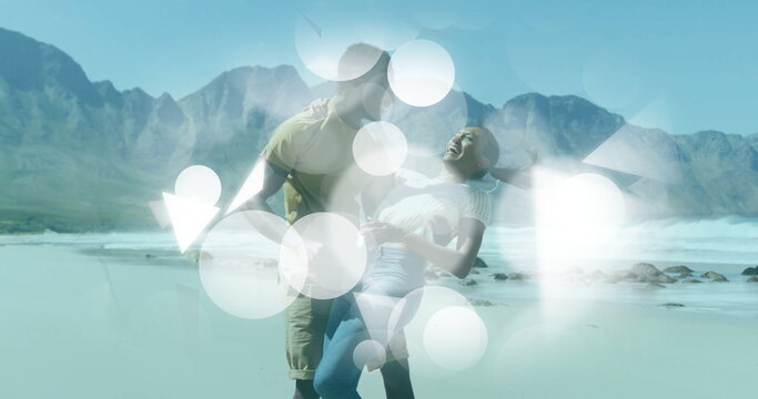 Romantic couple dipping and laughing on sunlit wet sand at beach shore, mountain peaks
