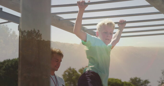 Climbing boy wearing green T-shirt on monkey bars at playground, supervisor wearing whistle lanyard - Powered by Adobe