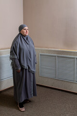 A Muslim woman in a modest gray hijab stands near a beige wall