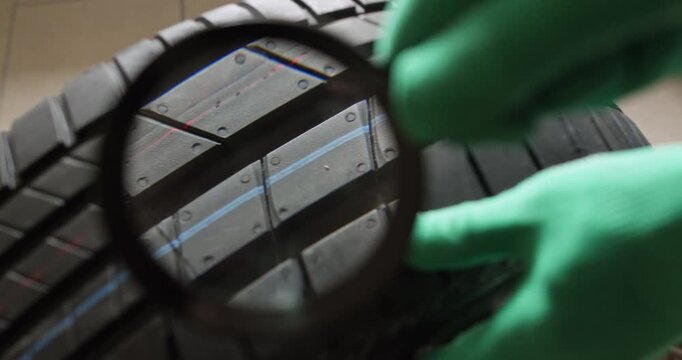 Mechanic in green gloves turns new car tire slowly while magnifier lens tracks rubber grooves. Gloved finger slides over tread showing block shape and tiny indicator
