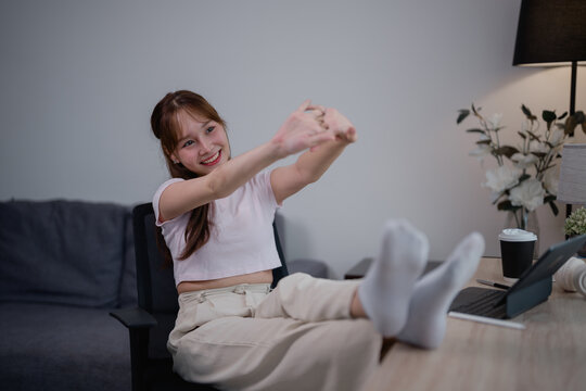 Freelancer woman is relaxing and stretching her arms while sitting on a chair with legs on the table in front of her tablet and keyboard after a long online remote work session