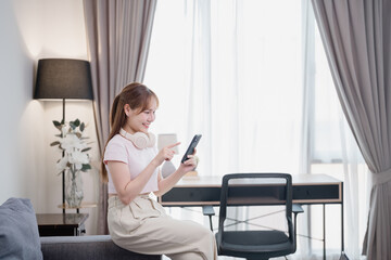 Happy young woman sitting on sofa and using mobile phone, wearing headphones around neck, in living...