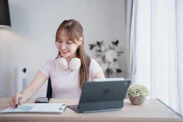 Young Asian businesswoman working from home sitting at her desk analyzing charts and taking notes,...