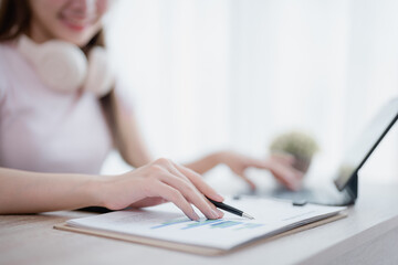 Asian businesswoman wearing headphones is analyzing financial charts with a pen in one hand while...
