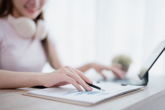 Asian businesswoman wearing headphones is analyzing financial charts with a pen in one hand while typing on a laptop with the other hand, working remotely from home - Powered by Adobe