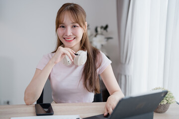 Smiling young asian woman working from home using tablet and headphones, sitting at her desk in a...