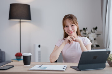 Young Asian businesswoman smiling while working from home, sitting at a modern desk with a tablet,...