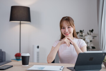 Smiling young Asian businesswoman working from home, using a tablet computer to manage tasks,...