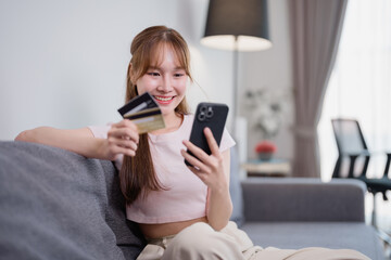 Smiling young Asian woman holding a credit card and using a smartphone for online payment while...