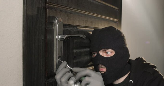 Man in black balaclava carefully inserts flathead screwdriver into lock of security door. Burglar smiles in satisfaction as screwdriver finally pops lock open