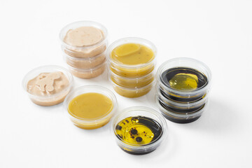 Assorted sauces in transparent plastic containers with lids on a white background. Includes light beige, yellow, and dark sauces (possibly soy or balsamic vinegar with oil)