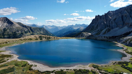 Fototapeta premium Serene alpine lake reflecting majestic mountain peaks under a vibrant blue sky with scattered clouds on a sunny day