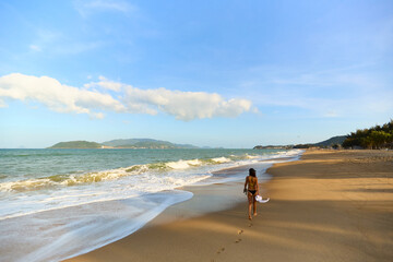 A solitary woman walks barefoot along a wide, sandy tropical beach at the water's edge, leaving footprints, with distant islands and mountains under a beautiful blue sky. 