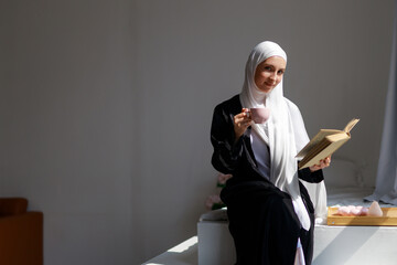 Portrait of beautiful young islamic woman in hijab drinking tea and reading book	
