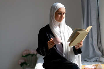 Portrait of beautiful young islamic woman in hijab drinking tea and reading book