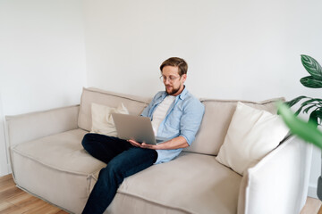 Man engages with laptop on sofa, immersed in remote tasks and highlighting balance of comfort and productivity enabled by digital tools in personal work routine.