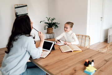 Mother and daughter share eye contact across tablet, creative markers on desk—capturing bond,...