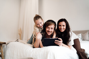 Fototapeta premium Man with headphones smiles while holding tablet as daughter and partner lean on him, symbol of digital family joy, screen interaction, and emotional bonding at home.