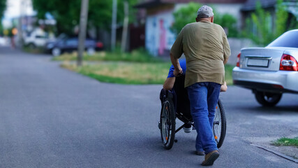 Obraz premium Elderly man pushes young person in wheelchair along suburban street during bright afternoon