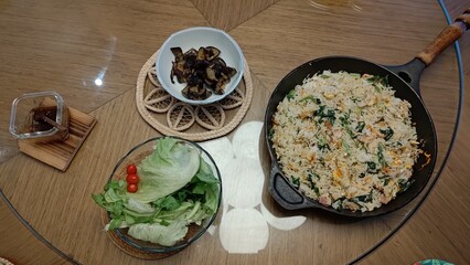 Top-down view of a complete homemade Asian meal featuring fried rice with vegetables and egg in a cast iron skillet, served with a side of fresh salad, roasted eggplant, and a dipping sauce.