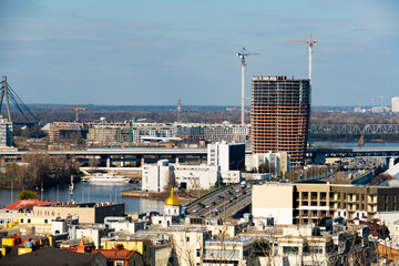 A panoramic view of the Kyiv cityscape featuring the Dnieper River, multiple bridges, modern buildings, and active construction sites under a bright blue sky