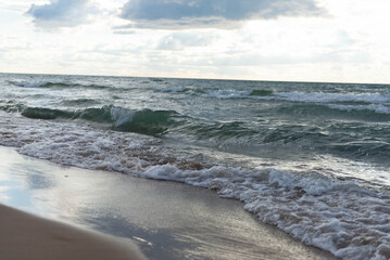 Close view of lake waves crashing on wet sandy beach. Stormy lake water washing onto wet sand with detailed texture of waves and foam. Natural scenic shoreline captured on Lake Michigan under sky.