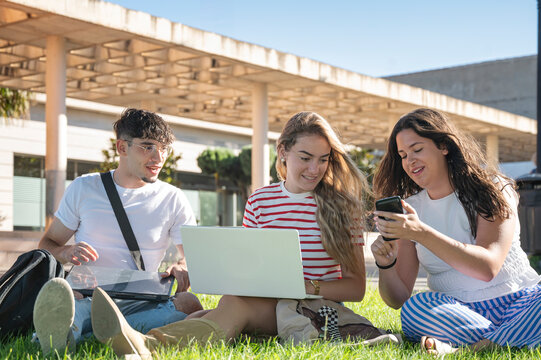 Three teenage students collaborating and sharing something on a smartphone while using laptops on the grass of a university campus during a bright sunny day of study and connection