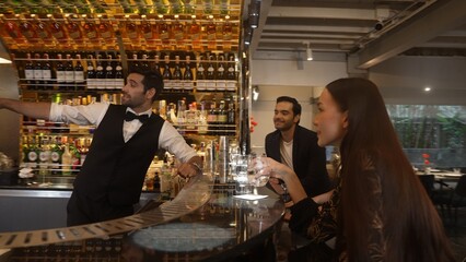 Smart bartender with VIP customer at luxurious bar greeting new beautiful guest at counter in engaging lively conversation at back view in lighting atmosphere at lighting warm nightclub. Vinosity.